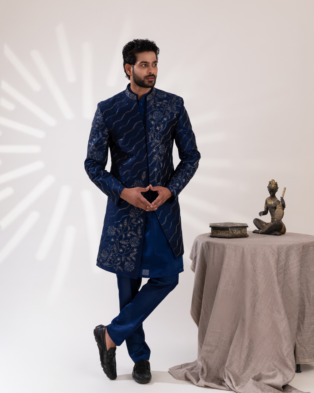 Man in a blue embroidered sherwani standing next to a table with a statue 