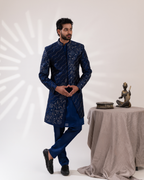 Man in a blue embroidered sherwani standing next to a table with a statue 
