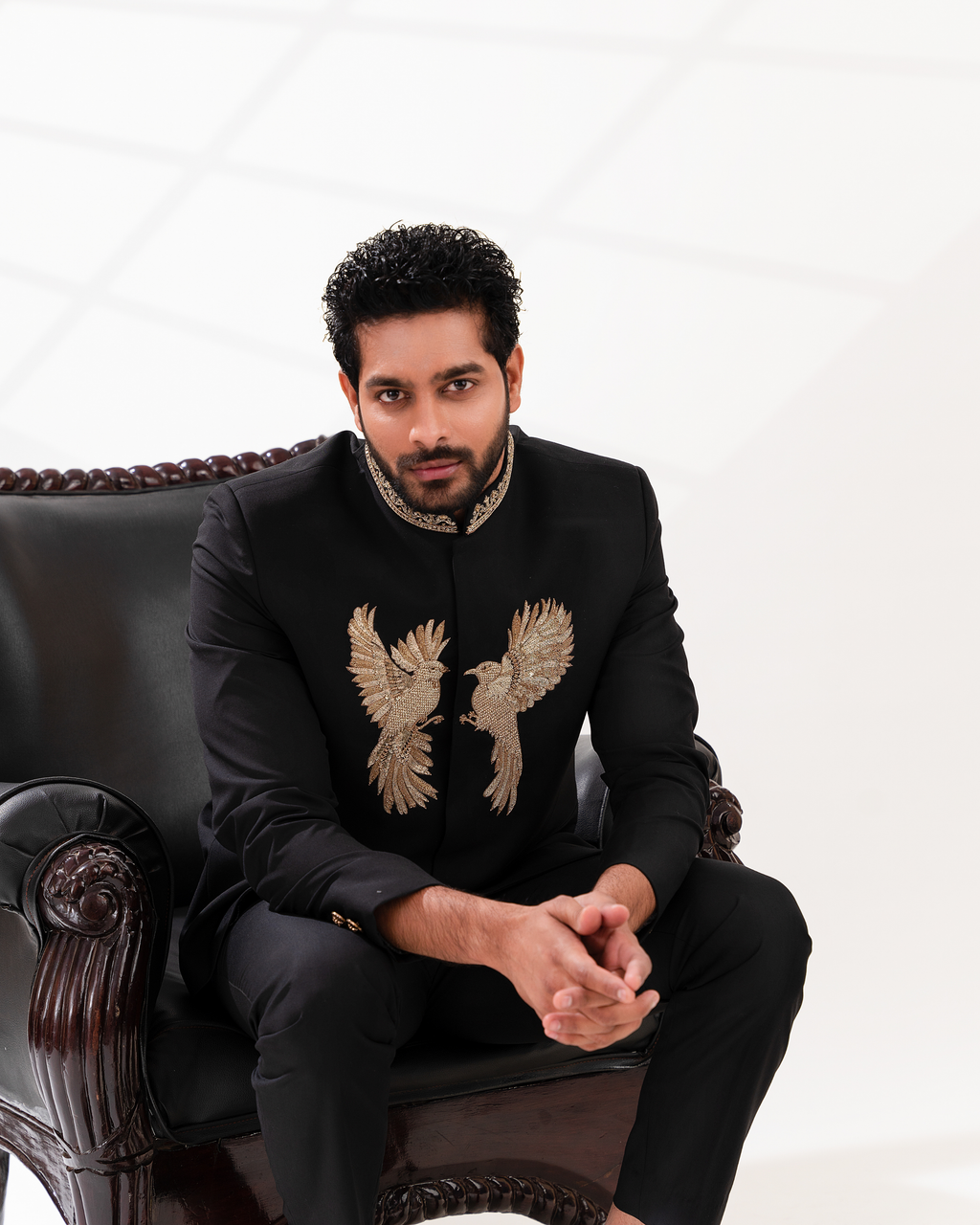 Man wearing a black suit with embroidered designs sitting on a decorative chair.