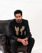 Man wearing a black suit with embroidered designs sitting on a decorative chair.