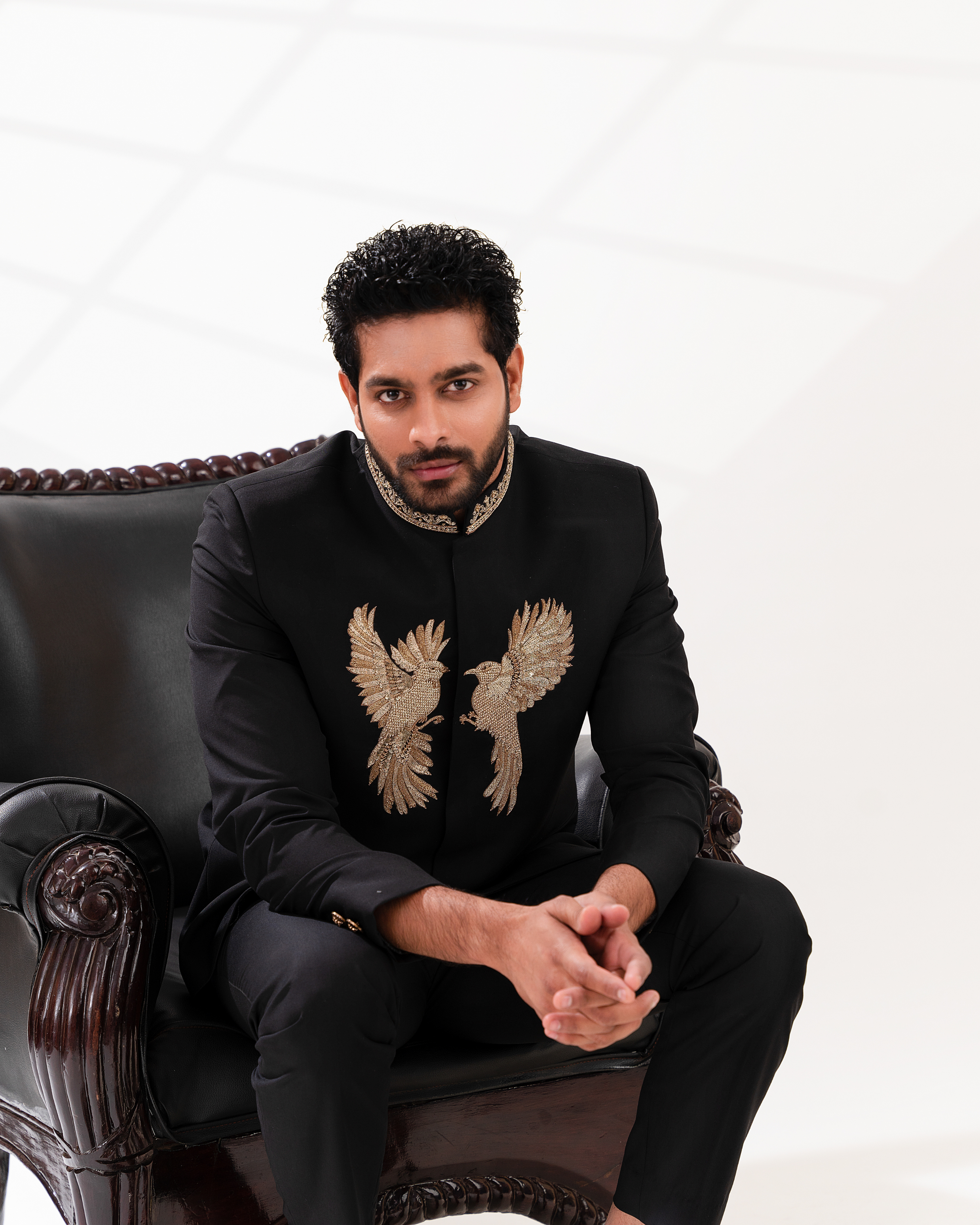Man wearing a black suit with embroidered designs sitting on a decorative chair.