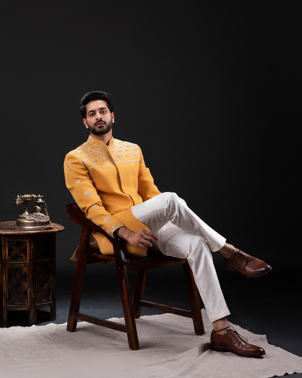 Man in a yellow embroidered bandhgala jacket and white pants sitting on a wooden chair